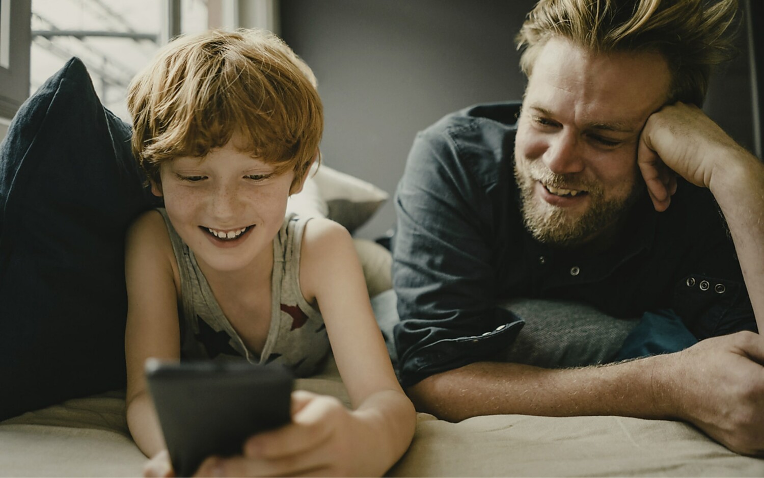 Vater und Sohn liegen auf dem Boden und schauen zusammen in ein Handy